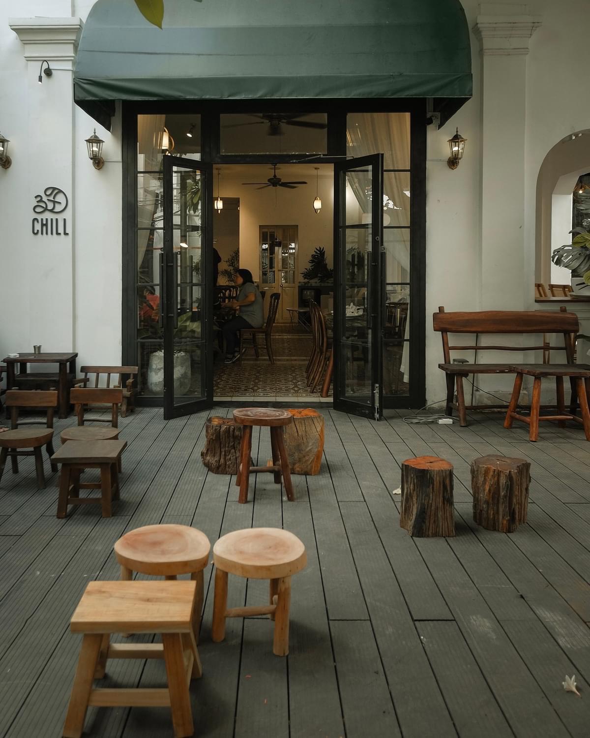 Sân trước quán ban ngày với ghế gốc cây và logo 3Đ CHILL trên tường trắng