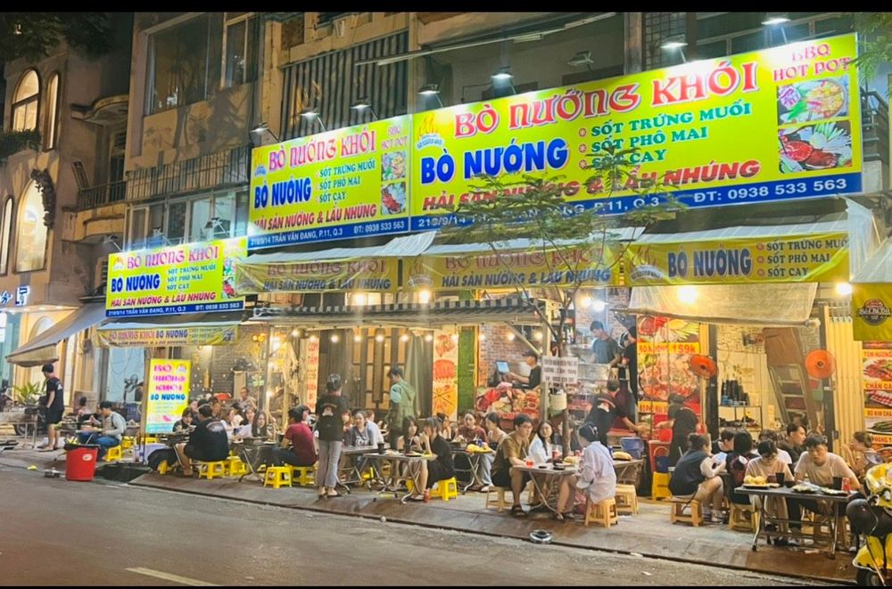 Mặt tiền quán buổi tối — bảng hiệu vàng, bàn vỉa hè