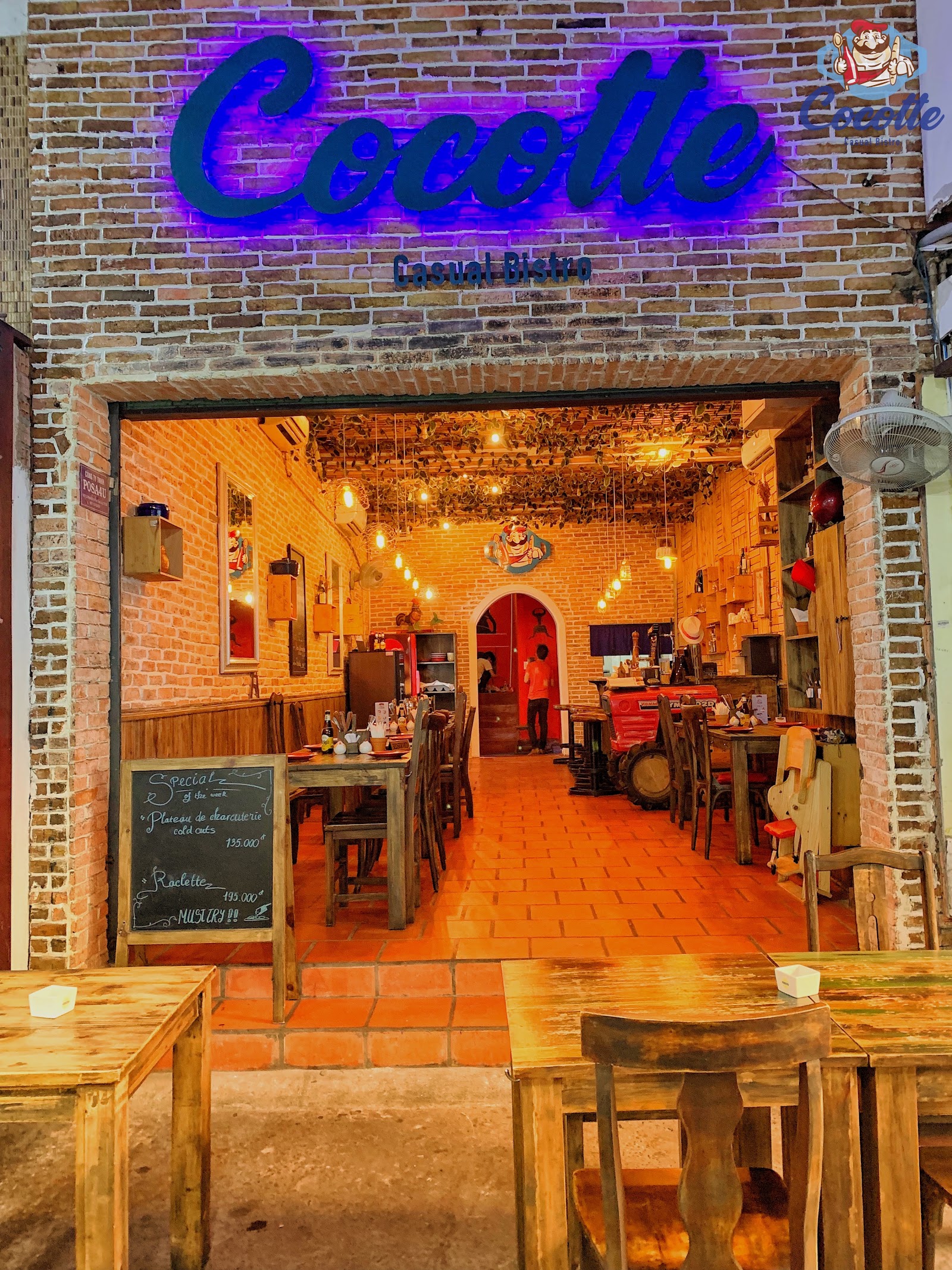 밤의 매장 전면: 벽돌 외벽, 파란 'Cocotte Casual Bistro' 네온 사인, 입구의 '이번 주 스페셜' 칠판, 앞의 나무 테이블
