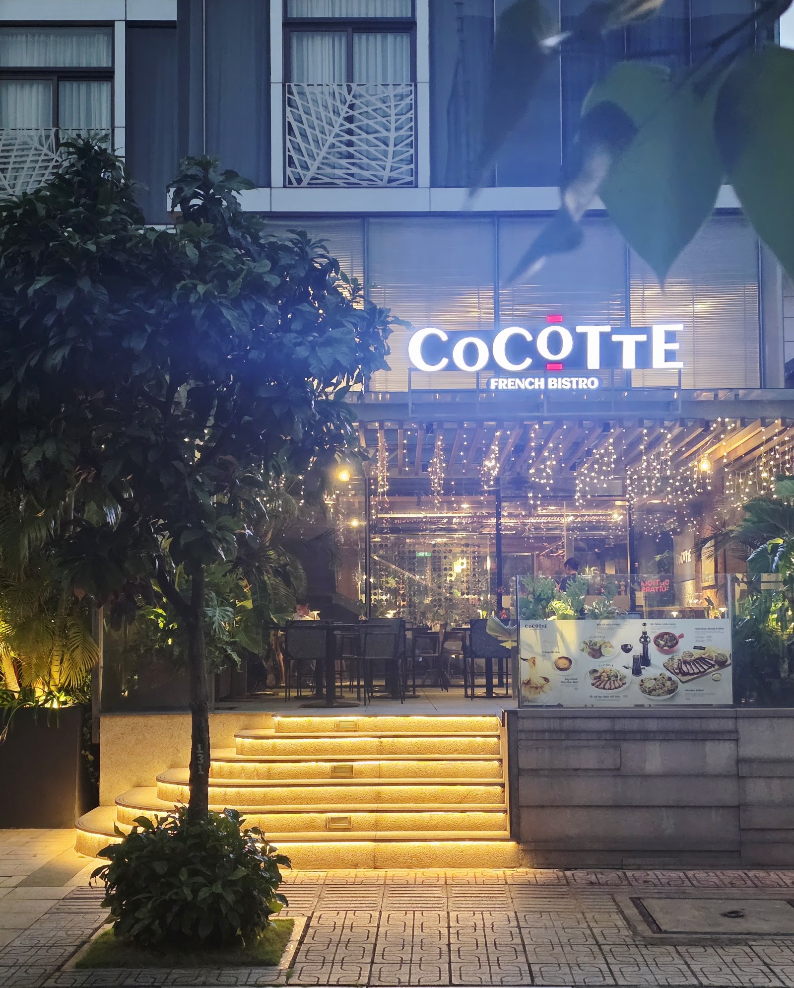 밤의 매장 전면: 테라스 위 빛나는 흰색 'CoCOTTE FRENCH BISTRO' 채널 문자, 따뜻하게 켜진 돌계단, 양옆의 나무