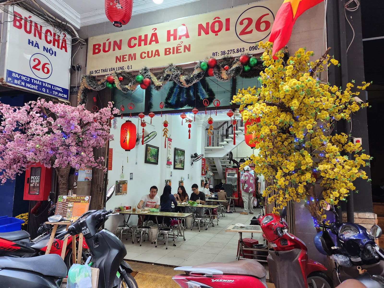 Mặt tiền ban ngày — thực khách ngồi ăn, cây đào hồng và cây mai vàng hai bên cửa