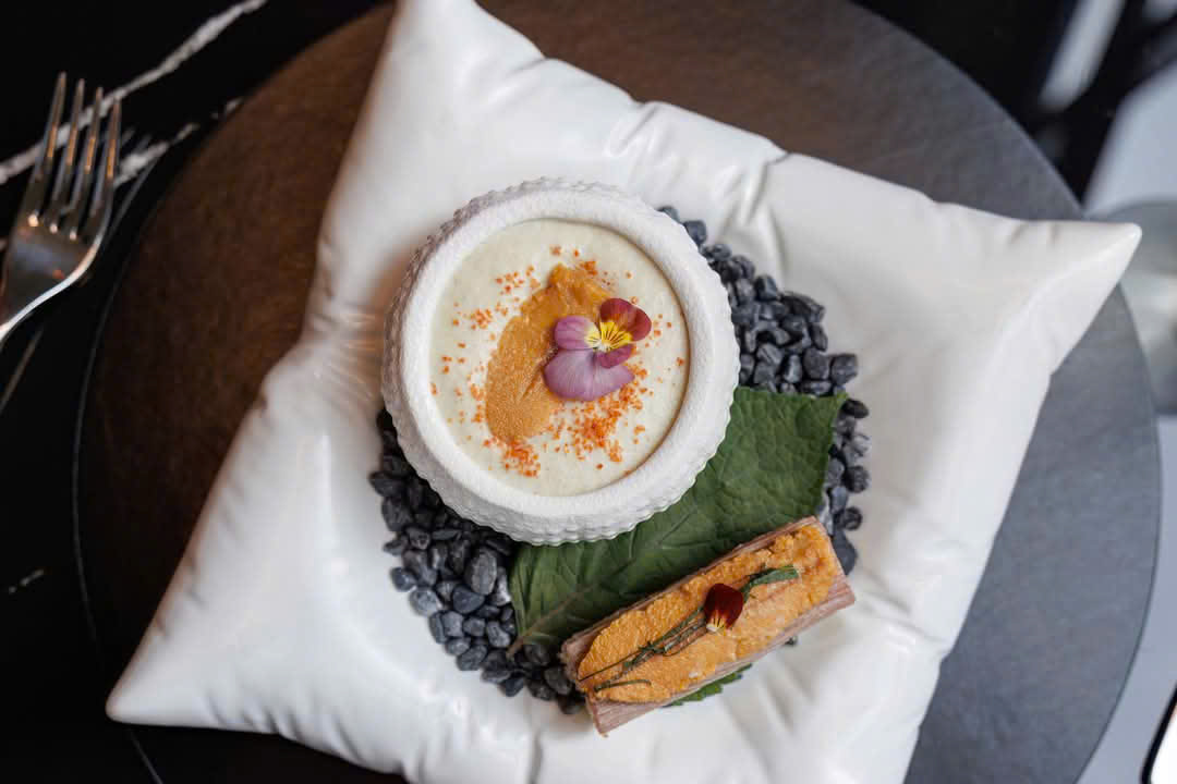 질감 있는 화이트 셸 볼의 성게 오버헤드 — 오렌지 우니·어란 가루·팬지 — 검은 조약돌·시소 잎·우니를 올린 프레스드 피쉬를 곁들인 베개 같은 화이트 도자기 위