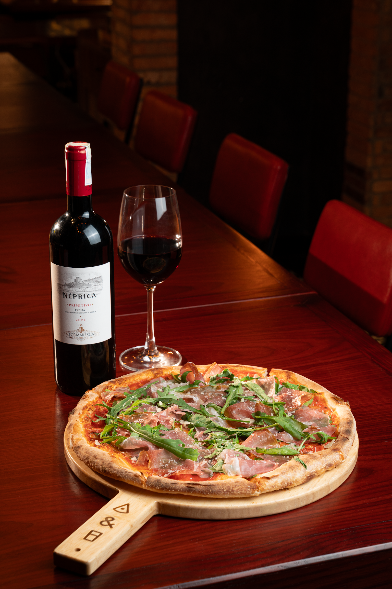 Pizza prosciutto và rocket trên khay gỗ cùng ly vang đỏ