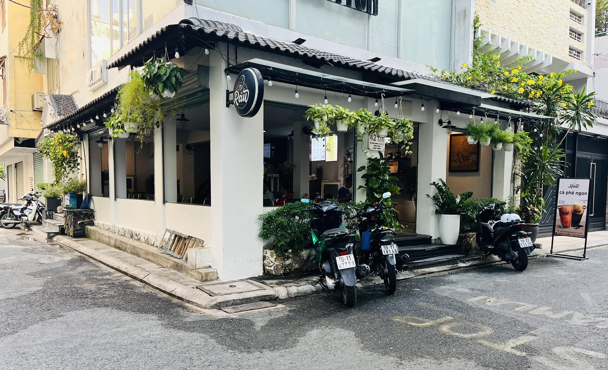 Râu Coffee 샵하우스 모퉁이: 둥근 검은 간판, 매달린 화분, 전구 줄