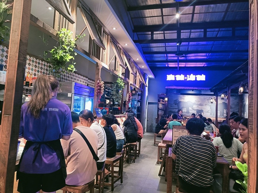 Bên trong Tiệm Ăn Siam: thực khách ngồi dọc quầy, tranh voi và bảng hiệu sáng ở cuối phòng